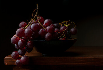 Baroque Renaissance still life in chiaroscuro style, with a bunch of red grapes in a black bowl on an antique wooden table with a dark background, decorative photography.