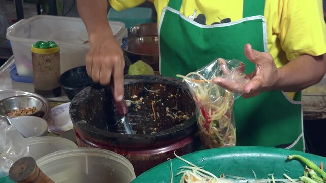 A street vendor skillfully prepares authentic som tam papaya salad, pounding fresh shredded papaya with chili, lime, fish sauce, and vegetables in a mortar for a flavorful Thai street food dish.