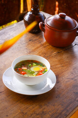 Traditional clear soup with boiled egg, fresh tomatoes, scallions, and vegetables served in a white bowl on rustic wooden table. Homemade Asian comfort food, healthy and warm meal.
