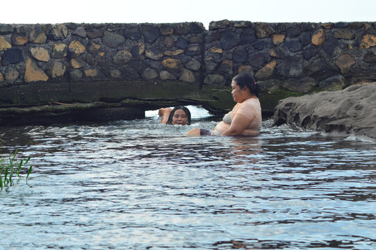 A mother and daughter enjoy playful relaxation in shallow brackish creek water near the sandy coastline, creating a peaceful yet joyful beachside atmosphere.