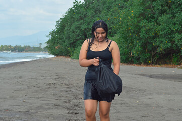 A girl pauses while walking on the sandy shore to check her black bag, smiling slightly, surrounded...