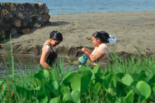 A mother and her teenage daughter sit close together in shallow water near the sandy beach, enjoying a calm moment by the riparian margin under natural sunlight.