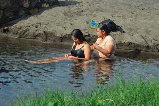 A mother and her daughter sit side by side in a shallow water channel near the sandy riverbank, preparing shampoo for a casual bathing moment in natural surroundings.
