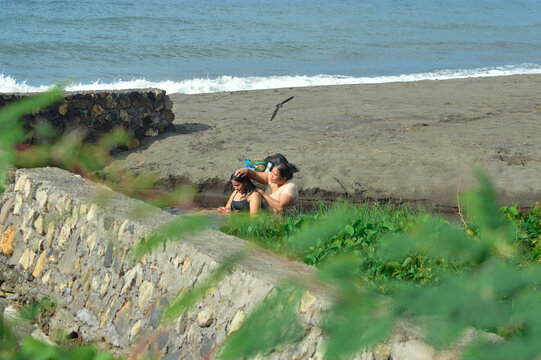 A mother washes her daughterâ€™s hair in a shallow water channel near the sandy beach while birds fly above, surrounded by green vegetation and bright coastal daylight.