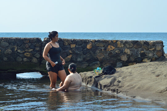 A teenage girl looks toward the horizon while carrying a soap bottle, standing in shallow brackish water as her mother sits close by near the sandy shoreline.