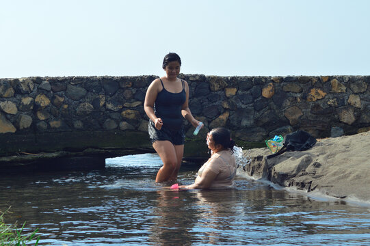 A mother splashes herself with water as her daughter walks nearby carrying a soap bottle, both in shallow coastal water near the sandy beach under bright daylight.