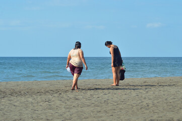 A teenage girl carries a small bag while walking with her mother on wet sand near foamy ocean waves, both enjoying the casual beachside atmosphere together.