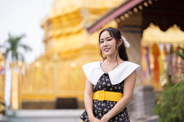 A young Asian woman wearing a stylish outfit with a yellow belt smiles confidently in front of a golden Buddhist temple