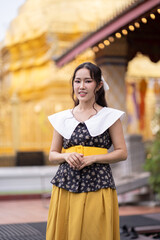 Asian woman in a retro-inspired outfit with a yellow belt poses with a gentle smile in front of a golden Buddhist temple.