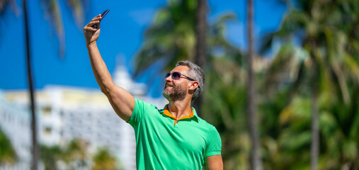 Tourist takes a selfie near palm tree. Man captures a selfie during summer vacation. Traveler snaps...
