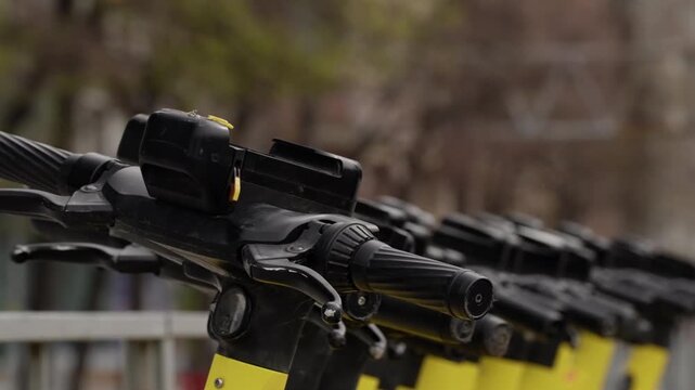 Close-up view of a row of yellow electric scooters along a city sidewalk, highlighting handlebars and controls for dockless micromobility and ecofriendly urban transportation