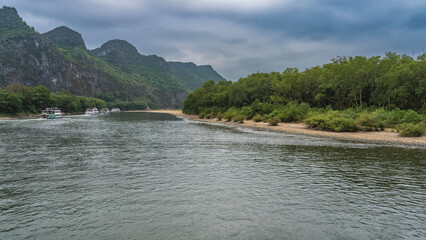 Tourist ships sail in the riverbed. Lush green vegetation on the banks of a calm river. Picturesque mountains against the sky and clouds. China. Li River. Li Jiang. Guilin