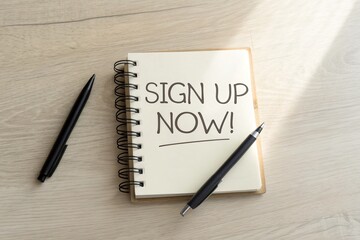 Open notebook on wooden table with a handwritten note urging to sign up now, surrounded by pens