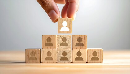 A hand places a wooden cube with a person icon on top of a pyramid structure made of similar cubes on a light wooden surface