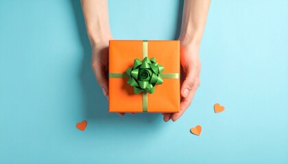 Aerial view of hands presenting an orange gift box tied with a green bow on a light blue backdrop, accompanied by three small orange hearts