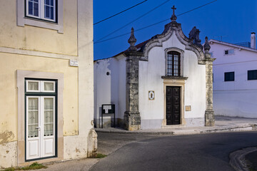 A small, ornate, white church building sits on a quiet street corner in Soudos, Portugal, with a traditional yellow and white building on the left.