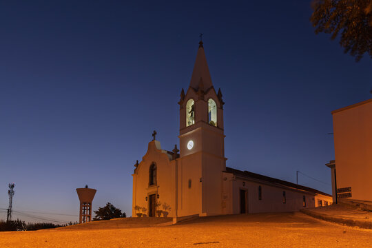 The Capela de Moreiras Grandes in Portugal, a charming white chapel with a bell tower and clock, is illuminated at dusk against a deep blue sky.