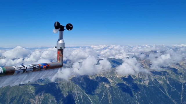​A close-up view of a wind speed sensor, or anemometer, positioned high on a mountain with a breathtaking panoramic view of a mountain range and a clear blue sky with puffy white clouds.