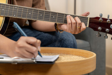 A creative young woman is enjoying her hobby, skillfully playing a guitar while concentrating on writing musical notes in her notebook. The scene captures the essence of music composition and artistic