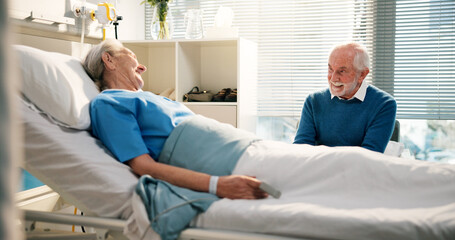 Obraz premium Talking, visit and senior woman in hospital with husband for bonding, support or care in recovery. Medical appointment, marriage and elderly female patient in ward bed for consultation in clinic.