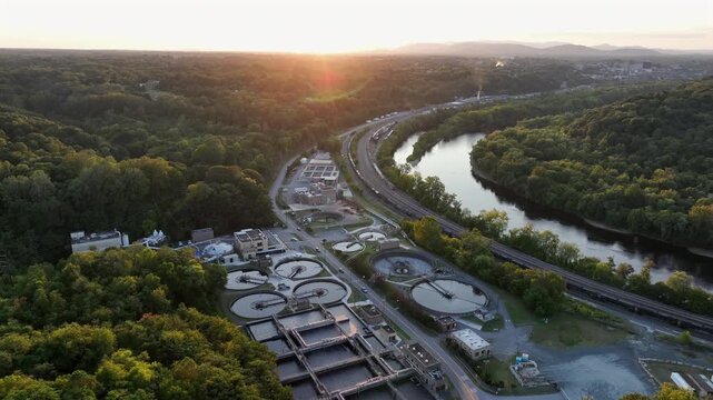 Aerial flyover wastewater treatment plant in American suburbia. Sunset time near James River with train on railways. Wide shot. Pools and ponds of sewage treatment plant in Virginia.