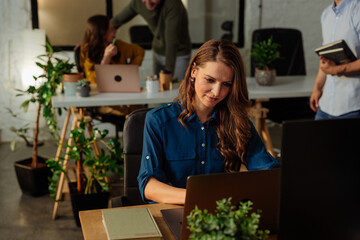 Focused businesswoman working on laptop in green office with colleagues talking in background