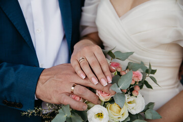 The bride in a white dress and the groom in a classic suit, holding a bouquet of roses, placed their hands on it, showing off their gold wedding rings