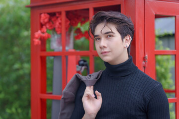 Young businessman in a black turtleneck holding a suit jacket and a briefcase, standing by a classic red telephone booth outdoors. Fashion portrait vintage style.