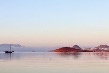 A peaceful sunrise or sunset over a calm, reflective sea, with sailboats and a small island in the distance, bathed in soft pastel light against a backdrop of mountains and a distant coastal town.