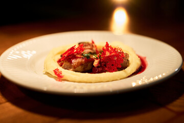 Gourmet dish featuring meat served on creamy mashed potatoes garnished with red decorative crisps presented on plate in restaurant setting with warm lighting