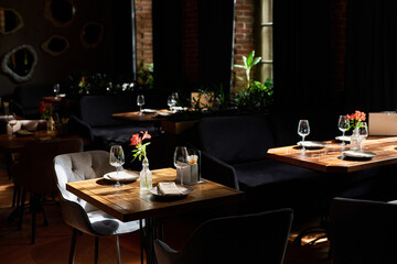 Empty restaurant interior showing wooden tables set with wine glasses, plates, cutlery and small flower vases, sunlight illuminating seating area near large windows, no people visible