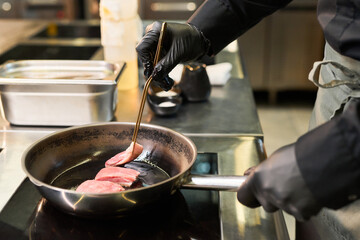 Caucasian man wearing black gloves cooking raw meat slices in frying pan using kitchen tongs, standing at stove in professional kitchen, focusing on precise food preparation