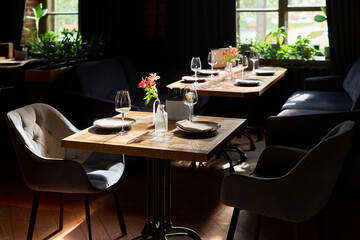 Empty restaurant tables with wine glasses, plates, cutlery, and floral centerpieces standing near large windows, sunlight illuminating wooden surfaces, modern dining setting