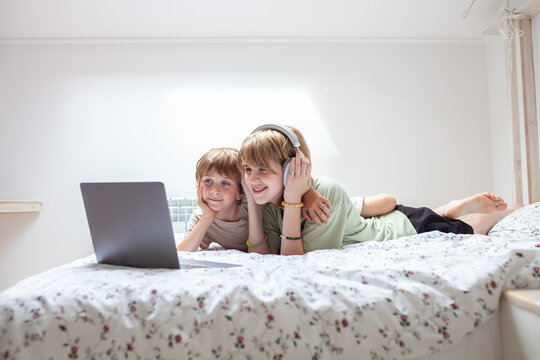 Two children lie on bed, looking at laptop screen with headphones and smiling. Listen to music, study with fun concept. Copy space. 
