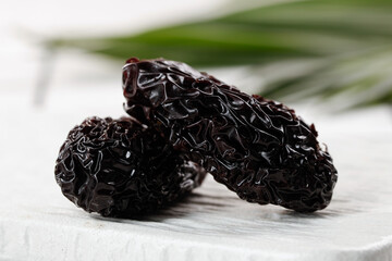 Black dried jujube fruits on white wooden base with shallow focus and natural palm leaf backdrop