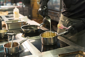 Caucasian young adult man wearing black gloves preparing food in professional kitchen, pouring cream into saucepan while stirring mixture of potato side dish on stovetop, cooking process in progress
