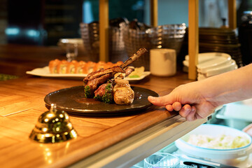 Caucasian young adult male hand serving plate with grilled lamb chops and vegetables on wooden counter in restaurant kitchen, sushi rolls visible in background, golden service bell nearby