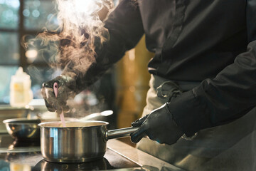 Middle aged Caucasian man wearing black gloves stirring food in stainless steel pot on stove, steam rising from pot, cooking in professional kitchen, torso and hands visible