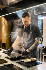 Caucasian young adult man with tattooed arm cooking food in professional kitchen, wearing apron and gloves, standing over stove and stirring ingredients in pan with steam rising