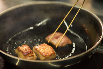 Unseen person using chopsticks turning pieces of meat while frying in pan on stovetop, steam rising from hot surface, close up showing cooking process in kitchen
