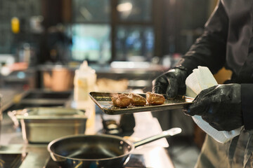Middle aged Caucasian man wearing black gloves holding metal tray with cooked meat pieces near stove in professional kitchen, preparing food with focus on hands and tray