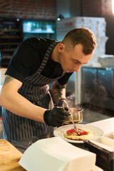Caucasian young adult man wearing apron and gloves carefully garnishing dessert with sauce in professional kitchen, focusing on precise plating technique for culinary presentation