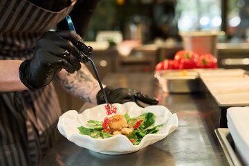Caucasian young adult man with tattooed arm and black gloves preparing gourmet dish in professional kitchen, arranging food with tongs on decorative plate, fresh vegetables in background