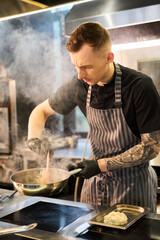 Caucasian young adult man with tattoos stirring food in pan on stove in professional kitchen, wearing apron and gloves, focused on cooking process, steam rising from pan