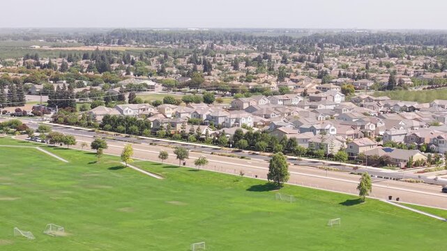 High drone shot receding above large soccer fields toward Ripon, California, showing road with light traffic and residential houses.