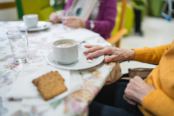 Elderly woman with wrinkled hand resting near coffee cup and two pills on saucer, having breakfast...