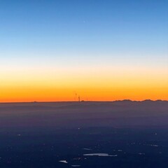 Dawn breaks; an orange horizon blends into a blue sky above hazy land marked by scattered lights and a distant tower silhouette