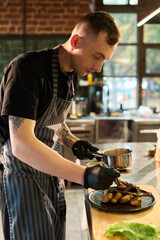 Caucasian young adult man with tattoos and short hair preparing gourmet dish in professional kitchen, wearing striped apron and black gloves, carefully plating cooked meat and vegetables