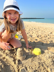 Happy little girl playing with sand toys on sunny beach, smiling child building sandcastle by sea, summer holiday fun outdoors