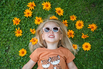 A happy little child girl in sunglasses lies on the grass surrounded by yellow flowers, top view,...